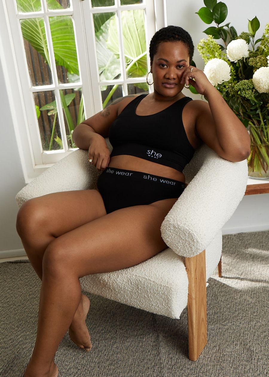 A woman seated by a window in a soft-cup black racerback crop top bra and organic bamboo G-string by an Australian company.