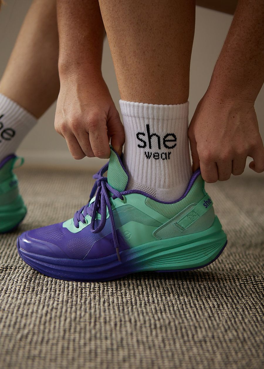 Woman adjusting her purple and aqua Australian-designed walking sneakers. She is wearing white cooldry sports socks.