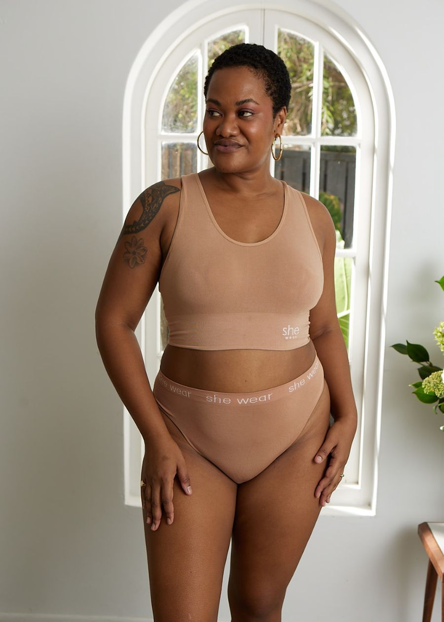 A woman near a window in a padded beige racerback crop top bra and G-string made of soft organic bamboo.