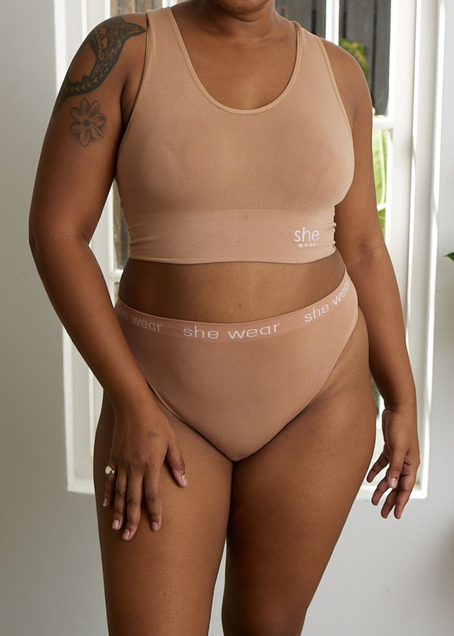 A woman by a window in a beige soft-cup black racerback crop top bra and organic bamboo G-string by an Australian company.