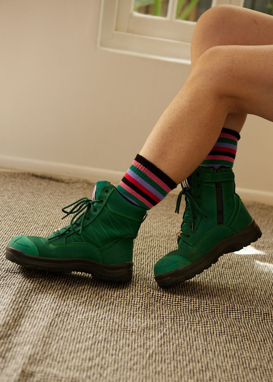 Indoor shot of a woman's legs in comfortable jade green safety cap boots with bright stripey organic bamboo work socks peeking out the top.